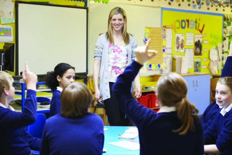 Teacher in a classroom with students raising their hands.