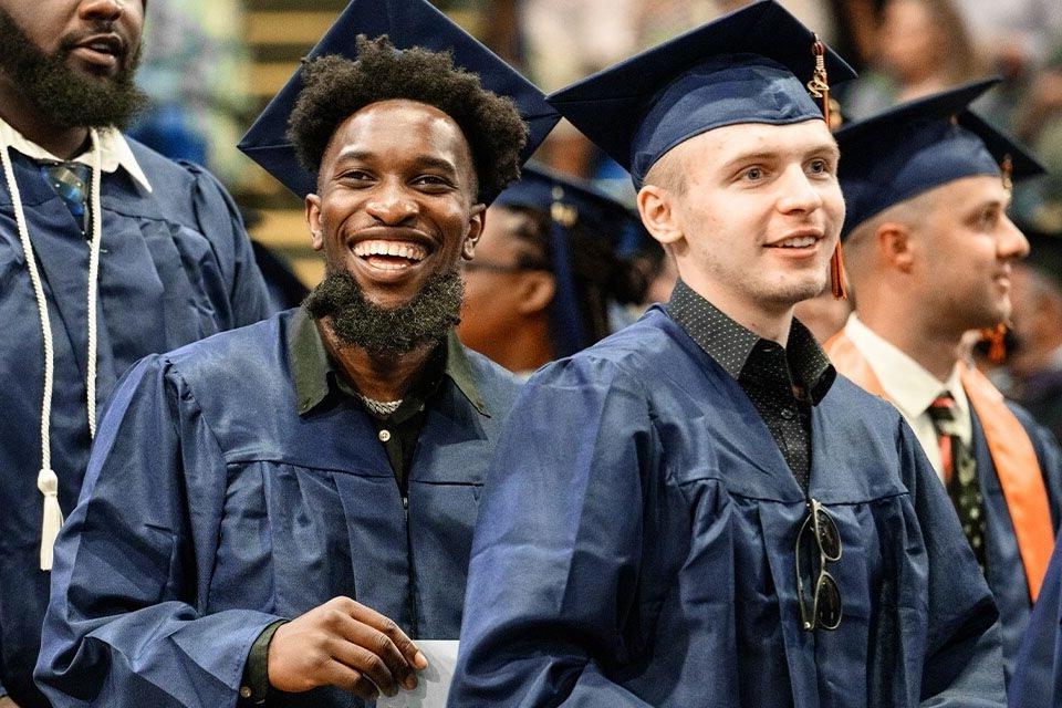 Three male Utica graduates smiling towards the crowed during graduation with blue cap and gowns on. 