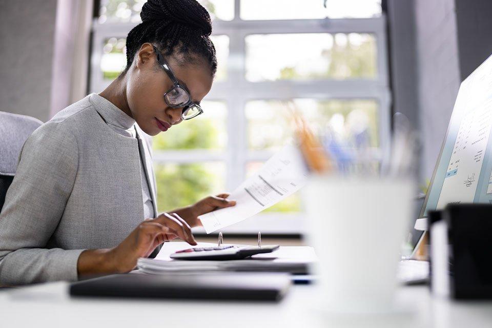 Black woman accountant reviewing paperwork and typing on a calculator in an office setting.